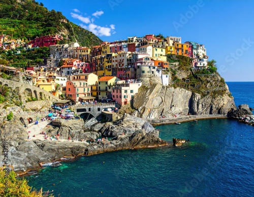Colorful houses cascade down a cliffside towards a vibrant blue sea. People gather on the rocky shore in this sunny coastal town landscape