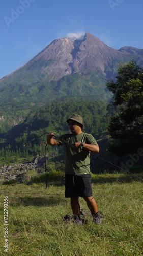 A male adventurer goes solo camping, setting up a tent in the grass with a beautiful volcano in the background. Outdoor activity: setting up a tent around an active volcano on a hot morning.