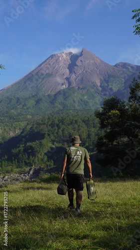 A male adventurer goes solo camping, setting up a tent in the grass with a beautiful volcano in the background. Outdoor activity: setting up a tent around an active volcano on a hot morning.