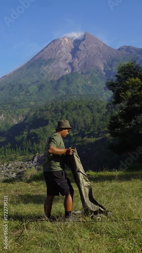 A male adventurer goes solo camping, setting up a tent in the grass with a beautiful volcano in the background. Outdoor activity: setting up a tent around an active volcano on a hot morning.