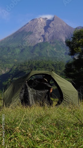 A male adventurer goes solo camping, setting up a tent in the grass with a beautiful volcano in the background. Outdoor activity: setting up a tent around an active volcano on a hot morning.