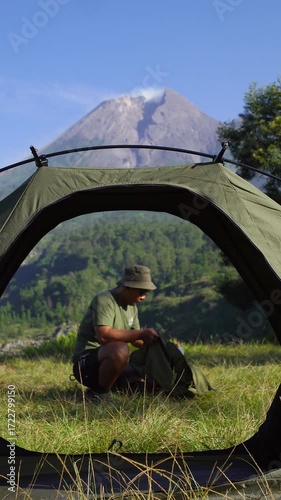 A male adventurer goes solo camping, setting up a tent in the grass with a beautiful volcano in the background. Outdoor activity: setting up a tent around an active volcano on a hot morning.
