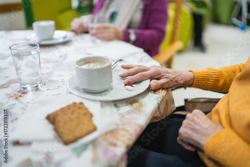 Elderly woman with wrinkled hand resting near coffee cup and two pills on saucer, having breakfast in a nursing home