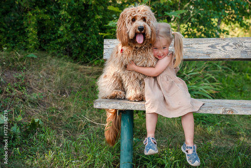 Canvastavla Little girl hugs a Curly brown goldendoodle or kawapoo dog in summer outdoor