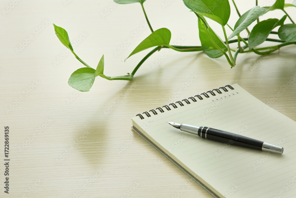 Fototapeta premium Notebook on wooden table, black pen, green plants, office supplies. Copy space