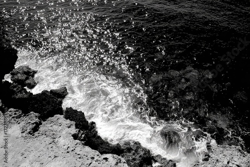 Black and white coastal scene. Waves crashing against dark rocks. Sunlight sparkles on water