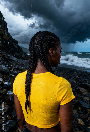A woman in a yellow shirt braids her hair while standing on a rocky beach under stormy skies.