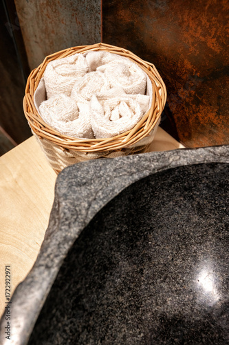 Close-up of basket with rolled towels next to stone sink on wooden counter. Spa-inspired bathroom detail with rustic tiles, natural textures, and clean wellness lifestyle concept.