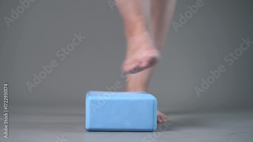 Individual balance training on a blue block with bare feet, demonstrating foot position and stability on a gray studio background