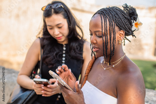 Multiracial women using smartphones outdoors. Two friends standing outdoors checking their phones.