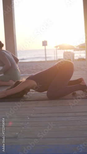 Pregnant woman with big belly doing yoga on the beach with trainer, woman lying in cat pose and pulling back.