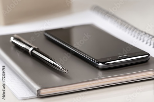 Phone rests on a notebook next to pen. Paper and spiral notebook in background. Light neutral office setting, close-up