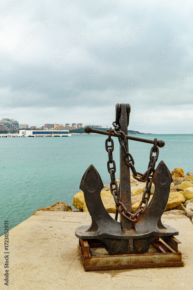 Fototapeta premium Rustic anchor stands on stone platform by the Black sea, with calm blue waters and cloudy sky creating peaceful coastal scene in Gelendzhik