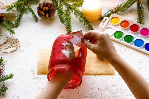 the boy's hands make a candlestick from a glass jar painted with red paint with a silhouette of a Christmas tree, step by step instructions, step 7.