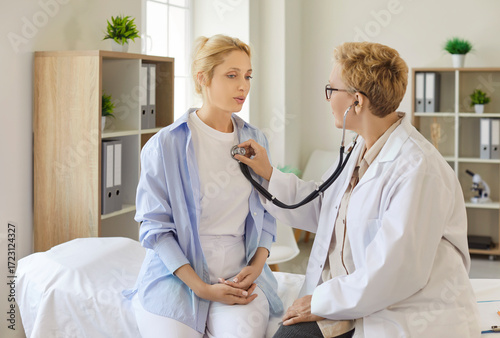 Fotografie Doctor using stethoscope to perform chest auscultation for young female patient sitting on exam bed in clinic and breathing