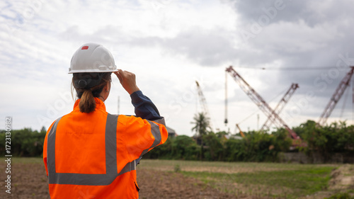 Foto A worker in an orange safety vest and helmet watches cranes at a construction site near a rural area