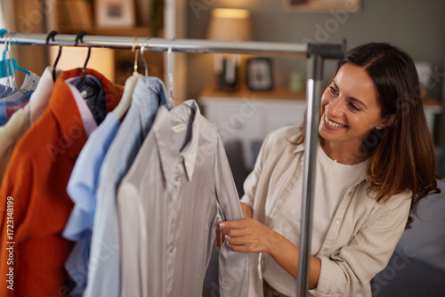 Murais de parede A woman is happily choosing shirts from a clothing rack in a bright and cozy room