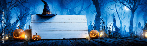 White Wooden Sign with Black Witch Hat and Glowing Orange Pumpkins in Dark Forest with Blue Moonlit Sky and Bare Trees in Background