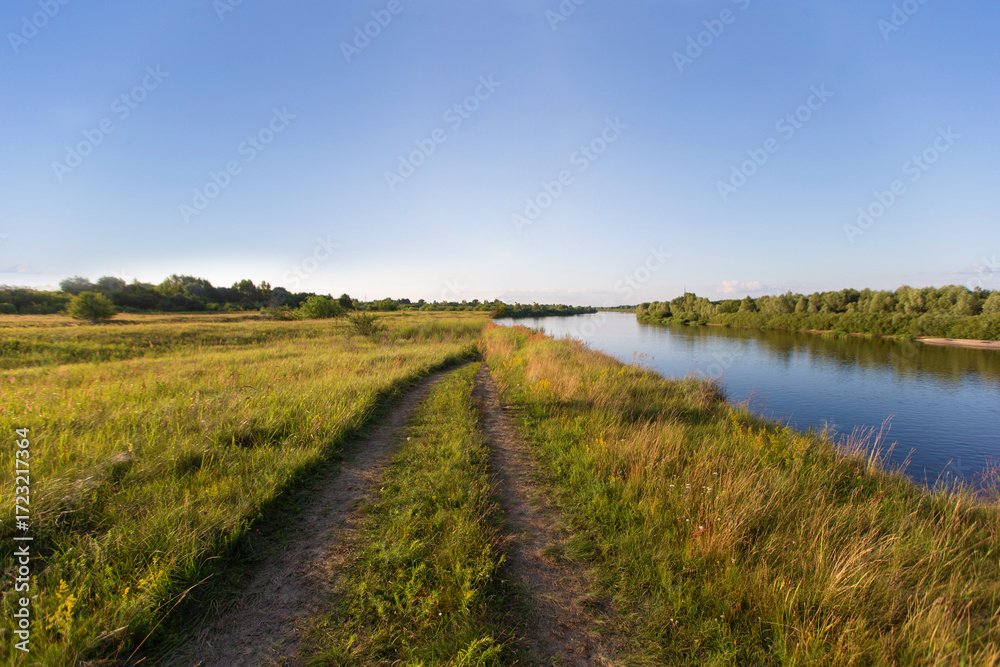 Obraz premium Exploring the serene riverside path surrounded by lush greenery on a sunny day near a gentle flowing river