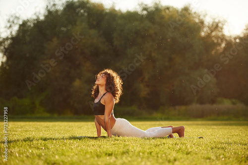 In black top and white pants, doing back stretching. Young fitness woman is on the field outdoors