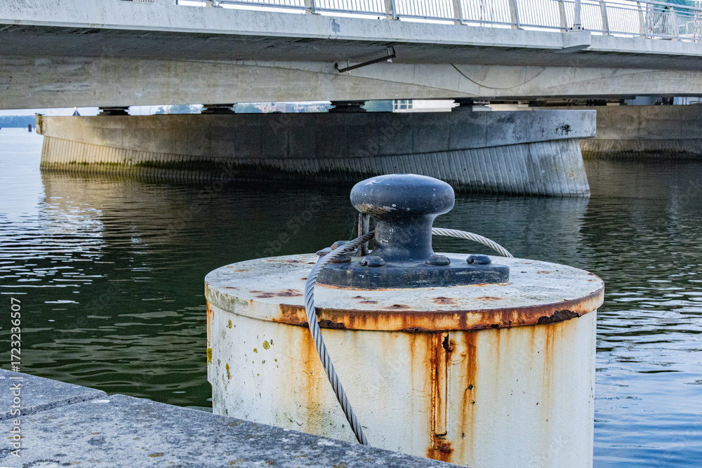 Fototapeta premium Close up of rusty mooring bollard with steel cable near concrete bridge over water