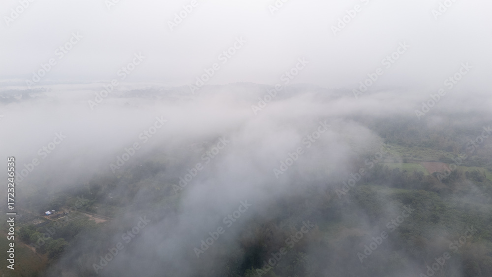 Fototapeta premium Aerial of Village in rain cloud cover tropical green mountain. Rainy season. Misty cover green forest. beautiful green village