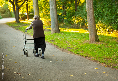 Elderly woman slowly taking a walk though park with help of a walker to aid 