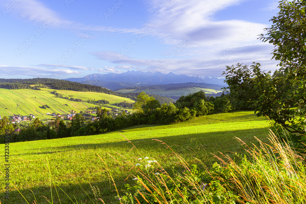 Obraz premium Sunlit hills with vibrant fields and scattered trees open toward distant blue mountains. A picturesque village nestles in the valley, embodying rural charm, traditional farming, and scenic tranquility