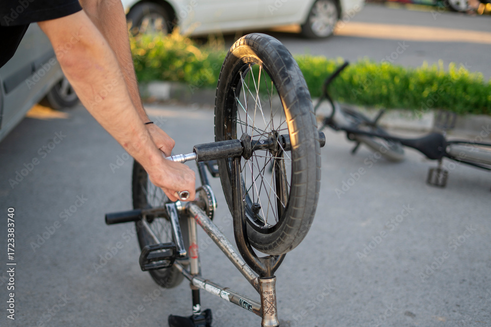 Obraz premium A person is focused on removing the back wheel of a BMX bike using a wrench, surrounded by parked vehicles in a well-lit parking lot.