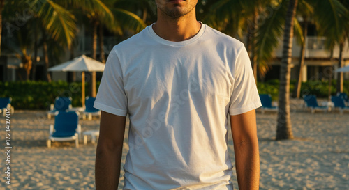 Man in plain white t-shirt standing on tropical beach with palm trees and lounge chairs at sunset
