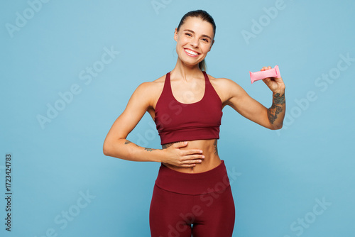 Young fitness trainer instructor woman sportsman wears red sportswear training in home gym hold energy protein bar put hand on belly isolated on plain blue background. Workout sport fit abs concept.