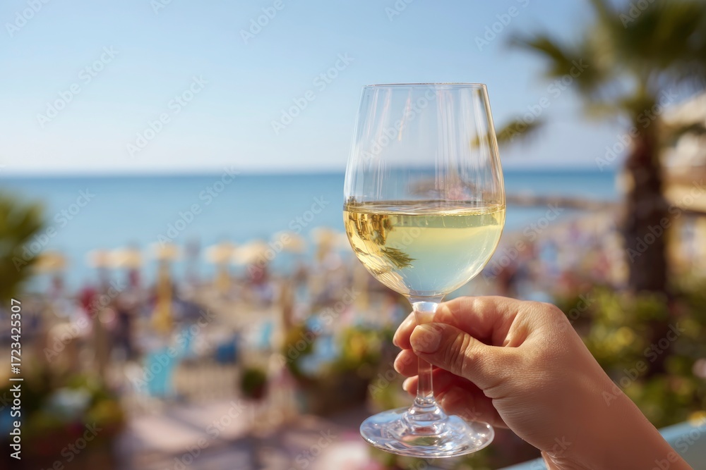 Obraz premium A hand holds a glass of white wine A beach scene with people umbrellas and palm trees is in the blurred background