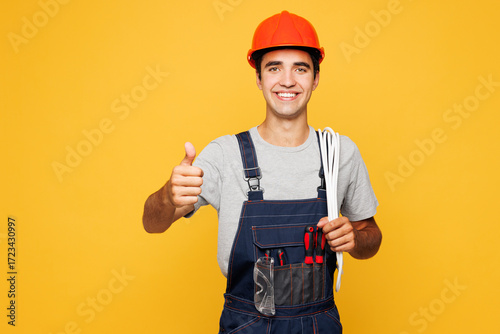 Young laborer handyman electrician man wear helmet overall carrying cables on shoulder show thumb up isolated on plain yellow background Instruments for renovation apartment room. Repair home concept