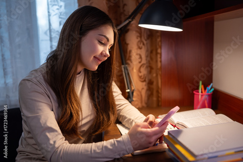 A female student got distracted by her phone while studying. A young girl, smiling, sits at a desk and gets distracted by her smartphone while doing her homework