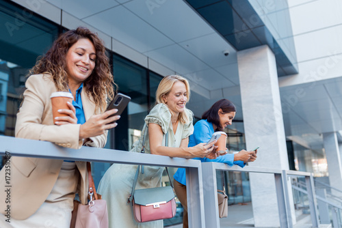 Group of businesswomen standing outside in front of modern office building looking at phone.