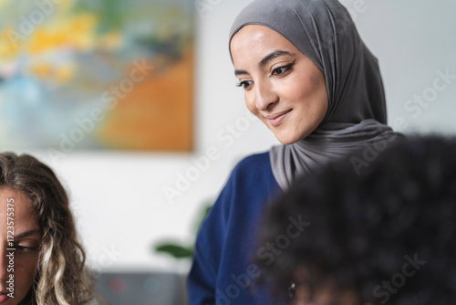 A young woman wearing a grey hijab and a blue top smiles softly, looking down. She is engaged in a conversation or activity with other people in an indoor environment.