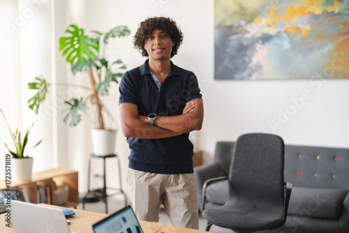A smiling young man with curly hair stands confidently with crossed arms in a modern office. He wears a navy polo shirt and light pants, looking directly at the camera.
