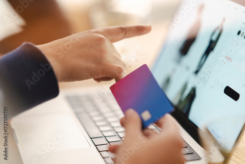 A person holds a credit card, pointing at a laptop screen displaying an online shopping website. They are actively engaged in adding items to a cart or completing an online payment