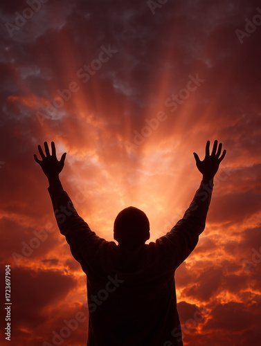 Rear view of a young man’s silhouette raising his hands at sunset, with warm red sunlight shining through him.