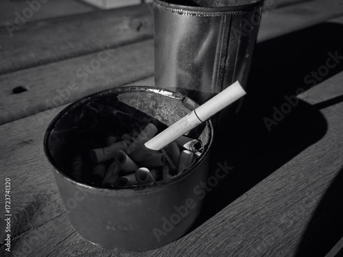 An ashtray was made from an old tin can, it is filled with cigarette butts, one cigarette is almost whole and smoking, a simple still life with a cigarette