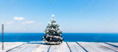 Geschmückter Weihnachtsbaum auf einem weissen Holzdeck vor einem Blauen Meer