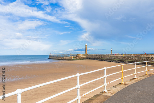 A pier with lighthouses stretches out to sea with a beach and railings in the foreground.
