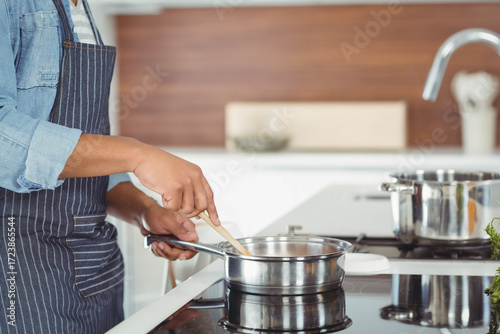 African American man in apron stirring skillet with spatula at cooktop in kitchen, copy space
