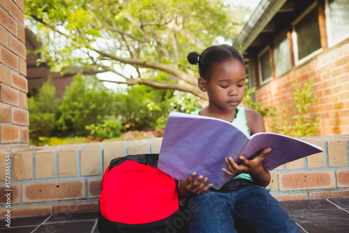 Child reading large purple book on low brick ledge outside building, with red and black backpack