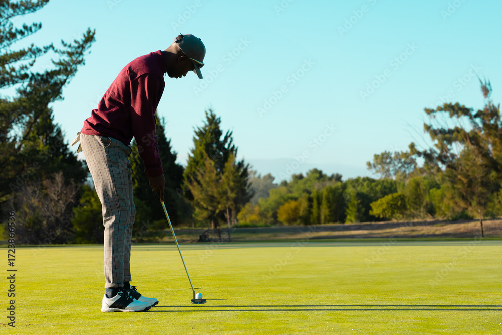 Obraz premium African American man lining up putt on putting green holding putter and ball, copy space