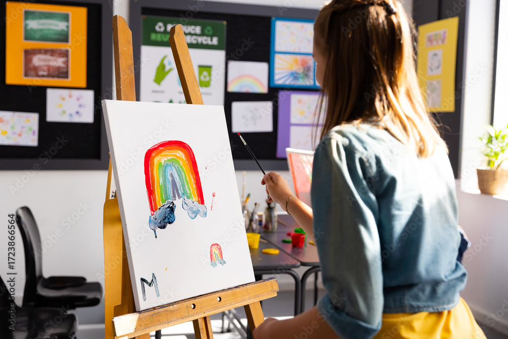 Naklejka premium School-age girl painting rainbow on canvas at wooden easel in art class with paintbrush, copy space