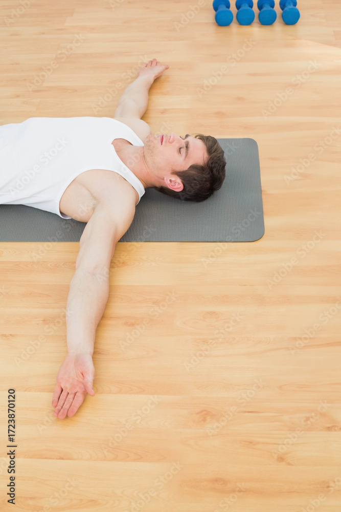 Fototapeta premium Man wearing white tank top and shorts lying on exercise mat in gym near blue dumbbells