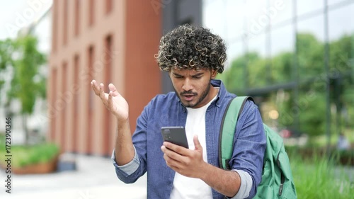 Worried male student read bad news on mobile phone while standing on street in campus space near university building. Frustrated stressed handsome man upset looking at message on smartphone. Close up