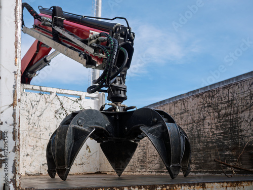 Industrial hydraulic grapple claw attachment inside truck container, showing black jaws, hoses, and rotator. Heavy-duty scrap metal handler used in recycling, salvage, demolition, and construction.