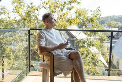 Casual man in white clothes sitting on balcony with coffee cup and smartphone, enjoying peaceful outdoor view of trees and houses. Concept of relaxation, modern lifestyle, and digital connection.
